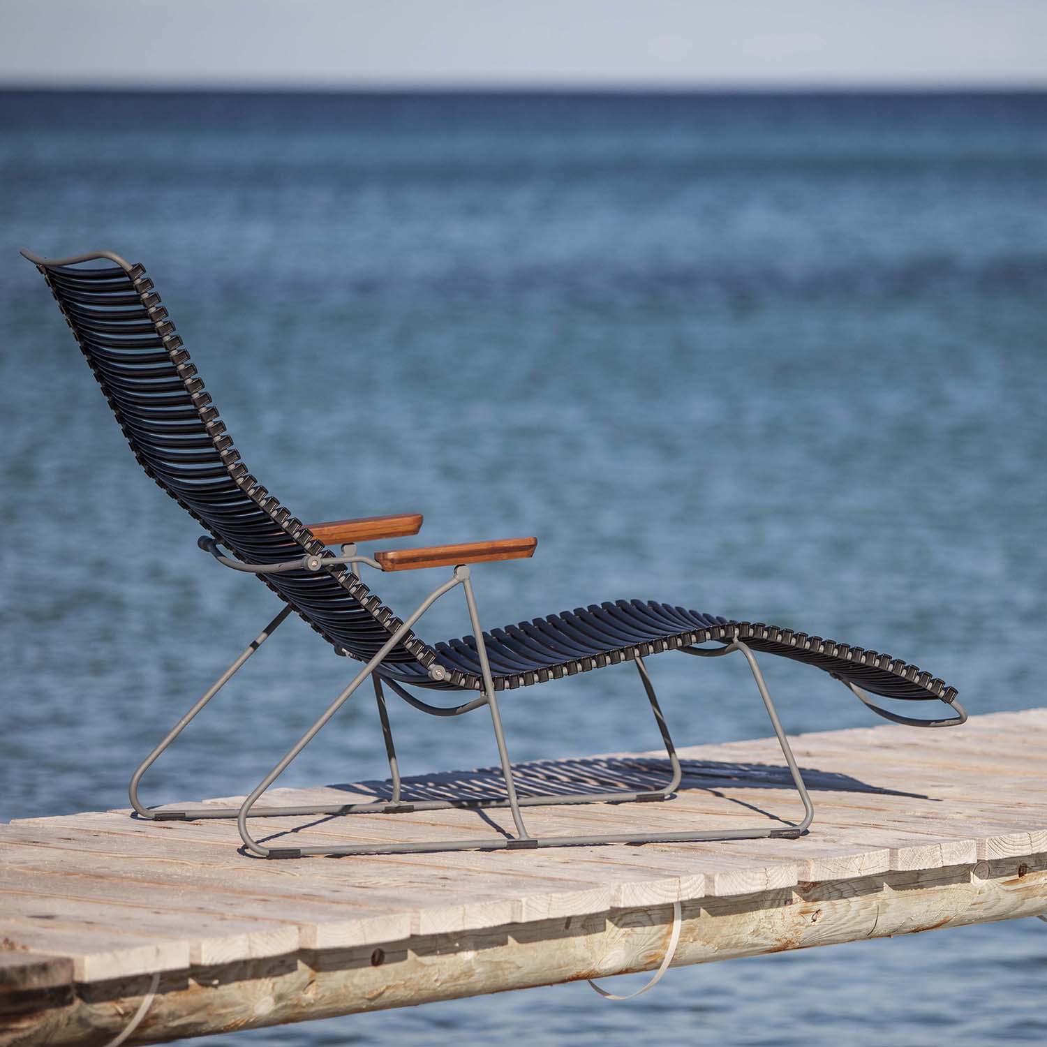 The Click Sunlounger by Houe, featuring stylish black design and wooden armrests for cushion-free seating, is set on a dock overlooking a tranquil blue sea. Its UV-protection makes it perfect for outdoor relaxation under a clear sky.