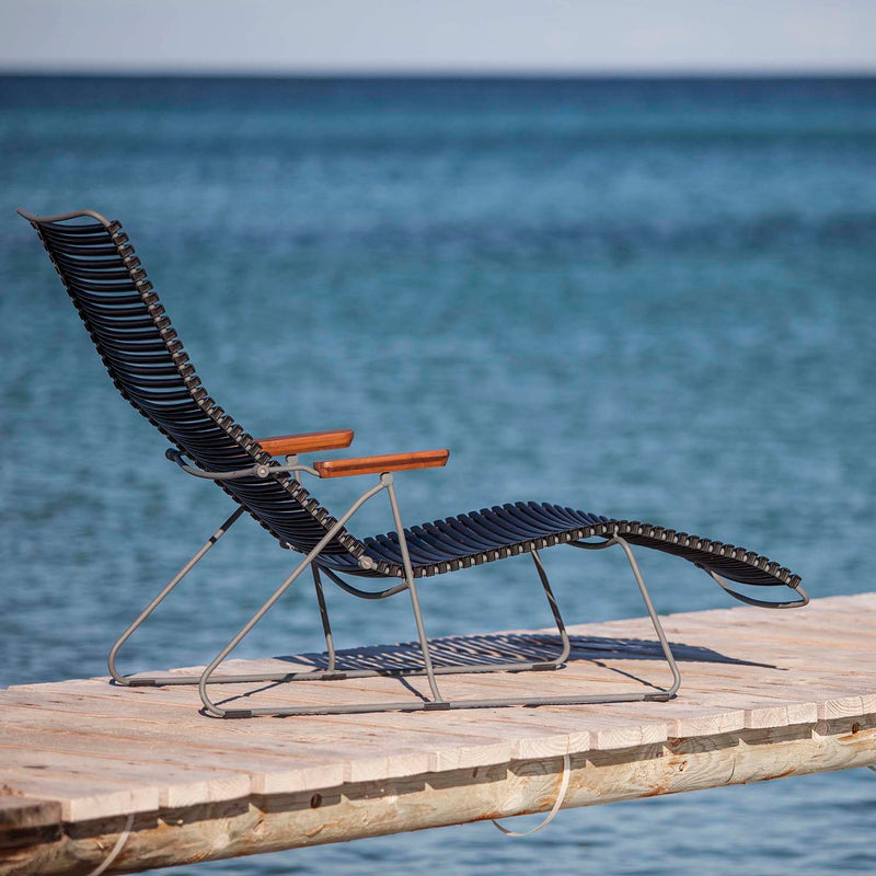 The Click Sunlounger by Houe, featuring stylish black design and wooden armrests for cushion-free seating, is set on a dock overlooking a tranquil blue sea. Its UV-protection makes it perfect for outdoor relaxation under a clear sky.