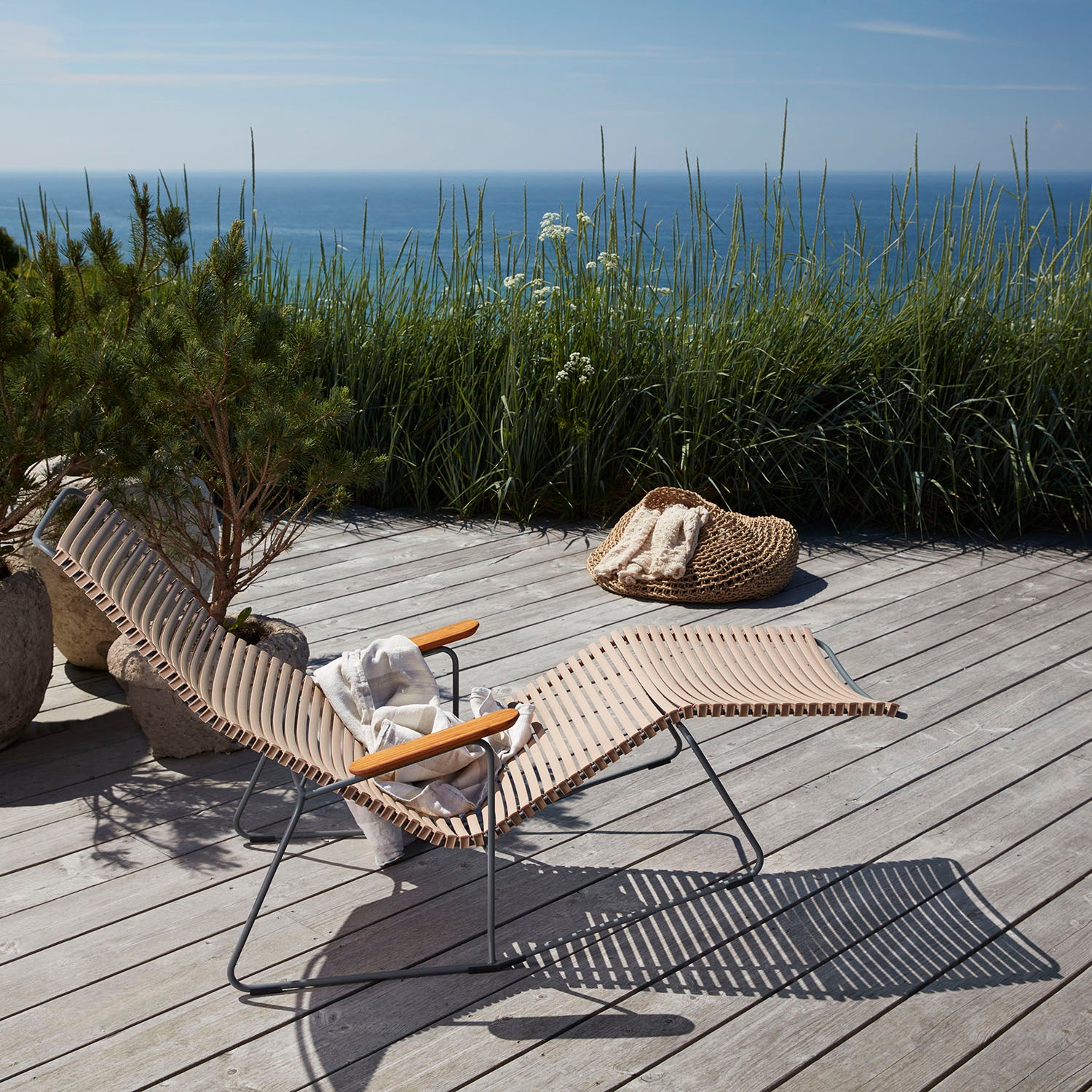A wooden deck overlooking the ocean features a Houe Click Sunrocker with bamboo parts and a striped design. A straw hat and bag rest on the rocker’s wooden armrests, surrounded by tall grass and potted plants under a clear sky.