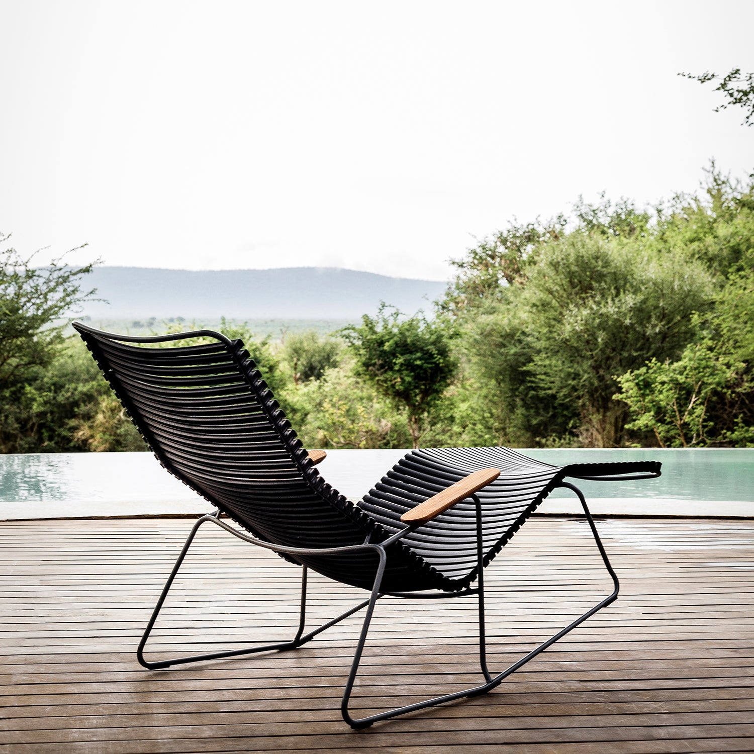 The Houe Click Sunrocker, a modern lounge chair featuring black design, wooden armrests, and bamboo components, rests on a deck overlooking a tranquil pool and lush greenery with distant hills under a cloudy sky.