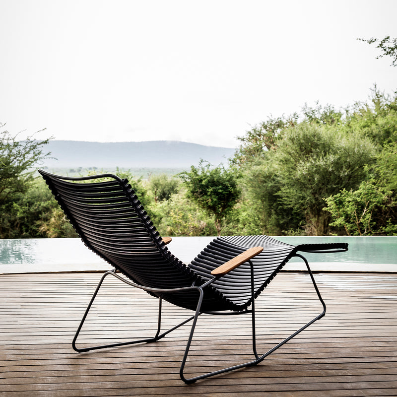 The Houe Click Sunrocker, a modern lounge chair featuring black design, wooden armrests, and bamboo components, rests on a deck overlooking a tranquil pool and lush greenery with distant hills under a cloudy sky.