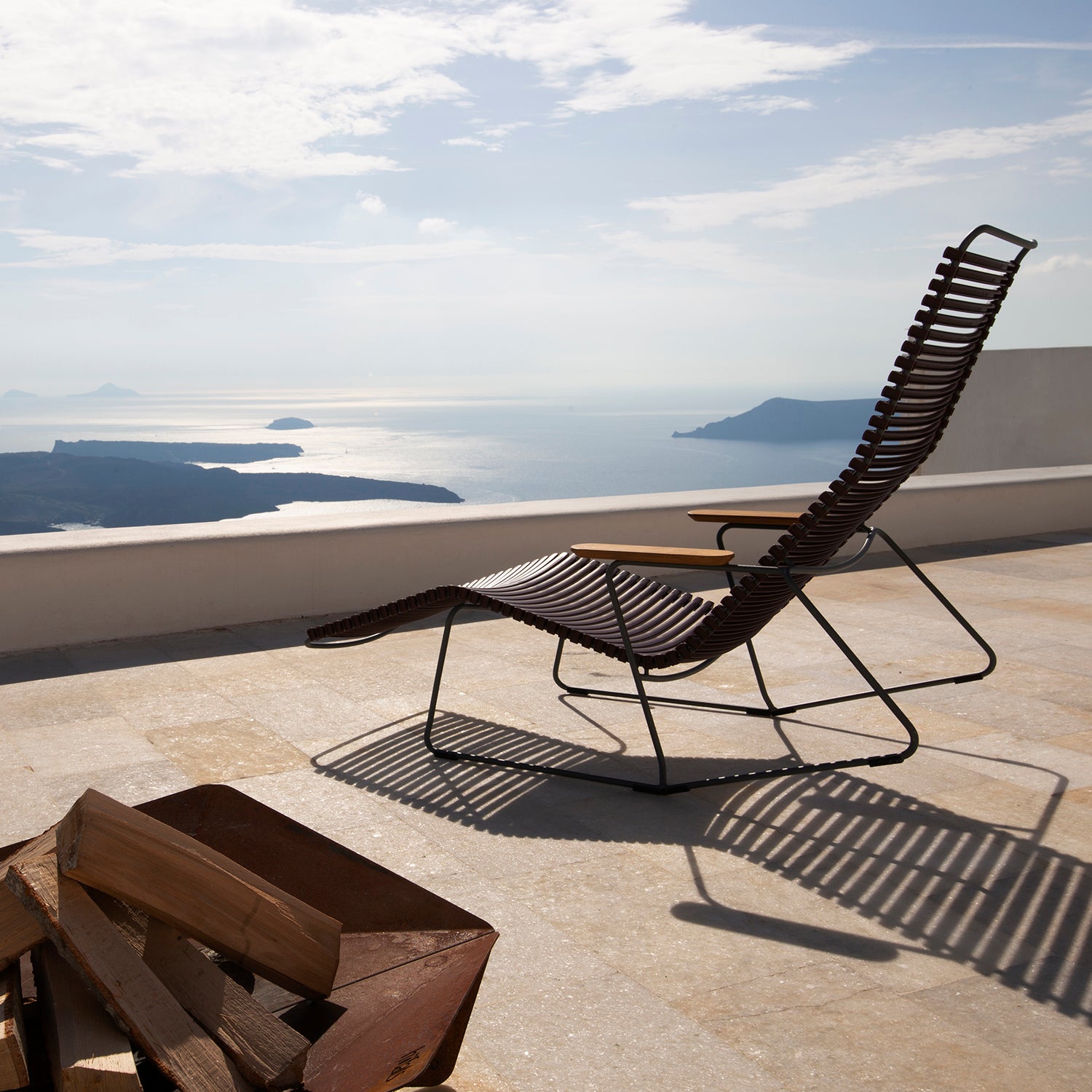The Houe Click Sunrocker, featuring a woven design and bamboo elements, is on a sunlit patio by the sea with views of distant islands. A clear sky enhances the serene ambiance, and a metal firewood holder is nearby.