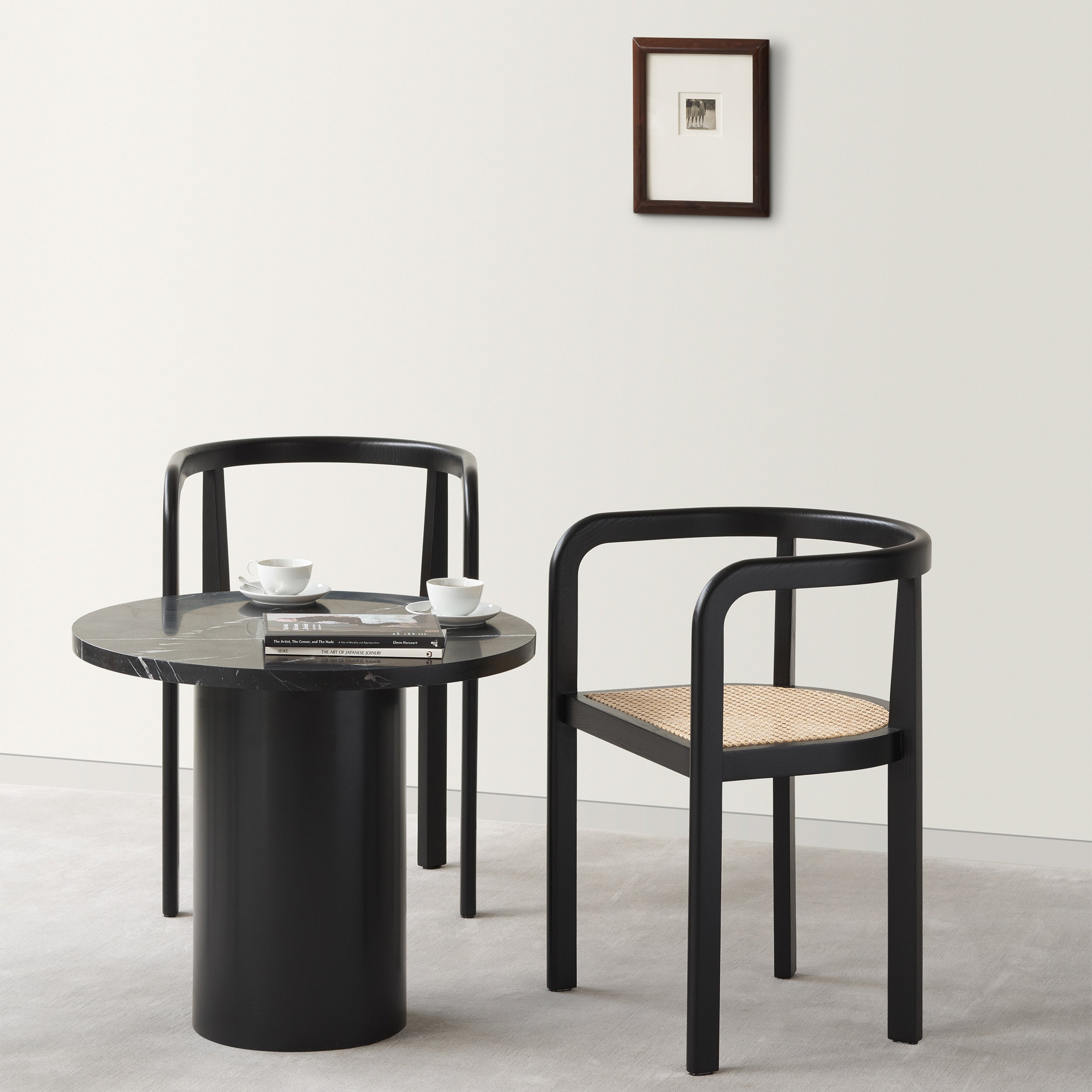A minimalist room showcases two black chairs and an E15 CT09 ENOKI BISTRO table with a rich marble top, holding two small white cups and books. A small framed picture adorns the white wall, while the light gray carpet complements the serene ambiance.