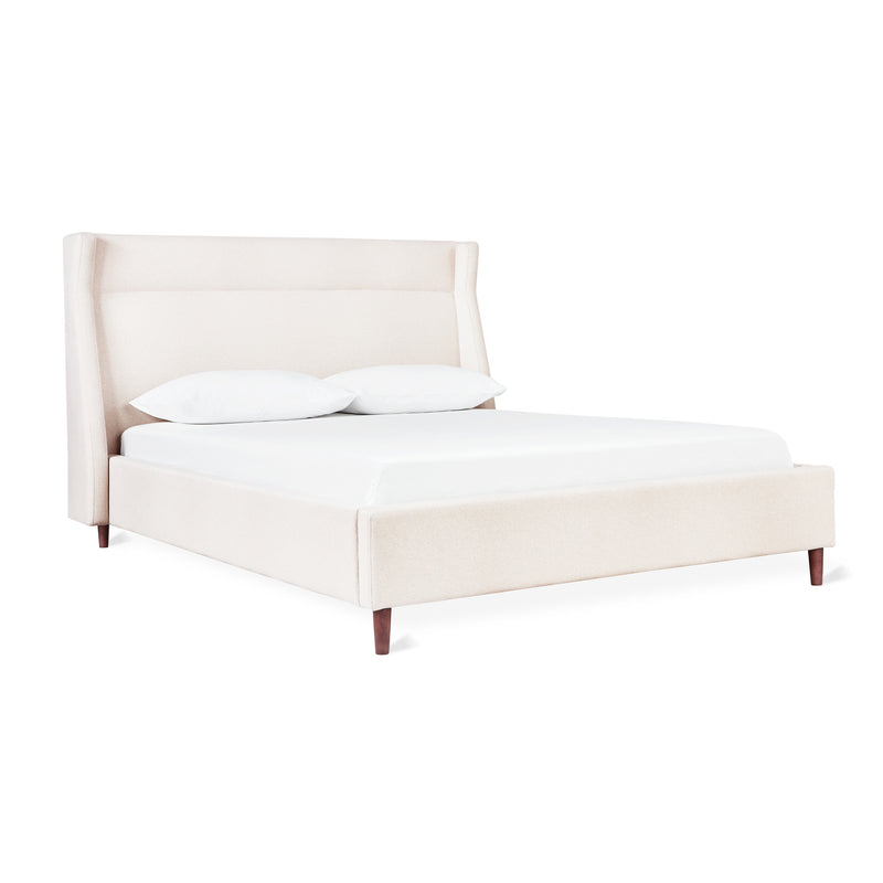 The Gus Modern Carmichael Bed features beige upholstery, an angled headboard, white bedding, two pillows, and wooden legs, all presented on a plain white background.