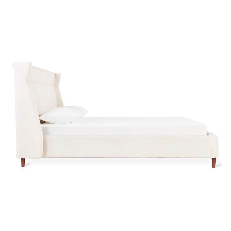 Side view of the Carmichael Bed by Gus Modern, an upholstered bed frame in off-white fabric featuring a tall wingback headboard, low footboard, and wooden legs, styled with a white mattress and single pillow.