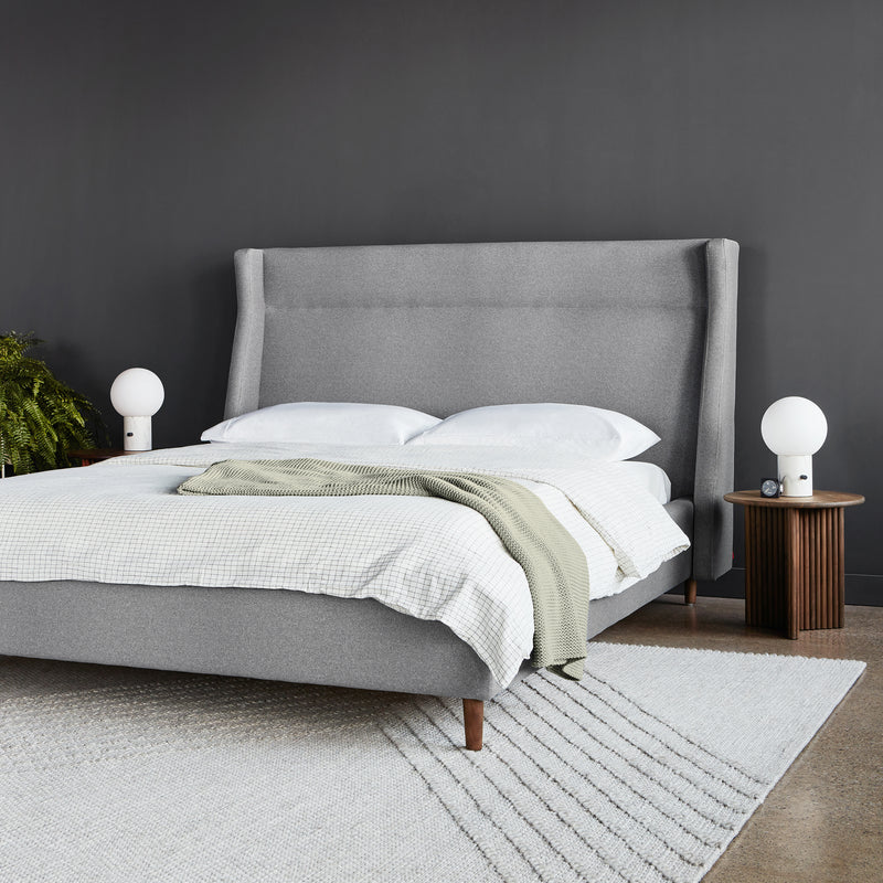 A modern bedroom with the elegant Gus Modern Carmichael Bed in gray upholstery, white bedding, and a green throw blanket. Wooden nightstands with white lamps flank the bed against a dark gray wall and light rug.