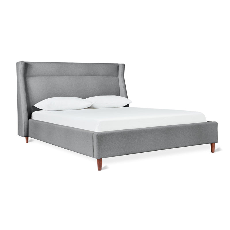 The Gus Modern Carmichael Bed features a tall, upholstered gray headboard, matching frame, wooden legs, and comes paired with a white mattress and two crisp white pillows.
