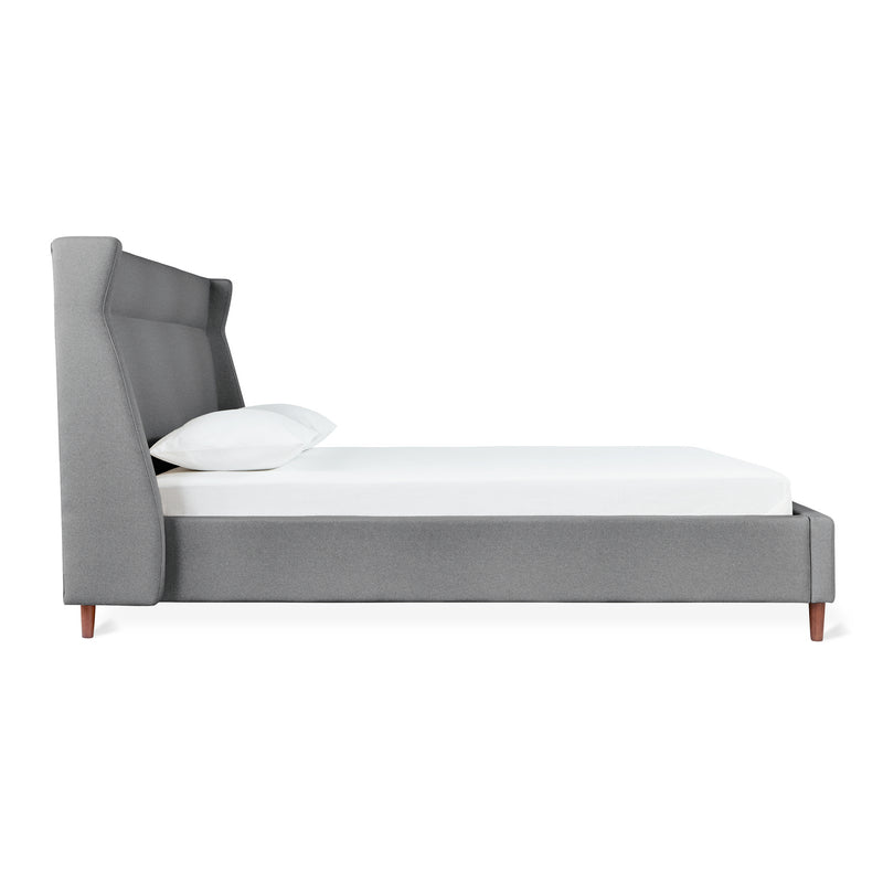 A side view of the Gus Modern Carmichael Bed, a modern upholstered bed with a tall wingback gray headboard, low-profile frame, wooden legs, white mattress, and a single white pillow.