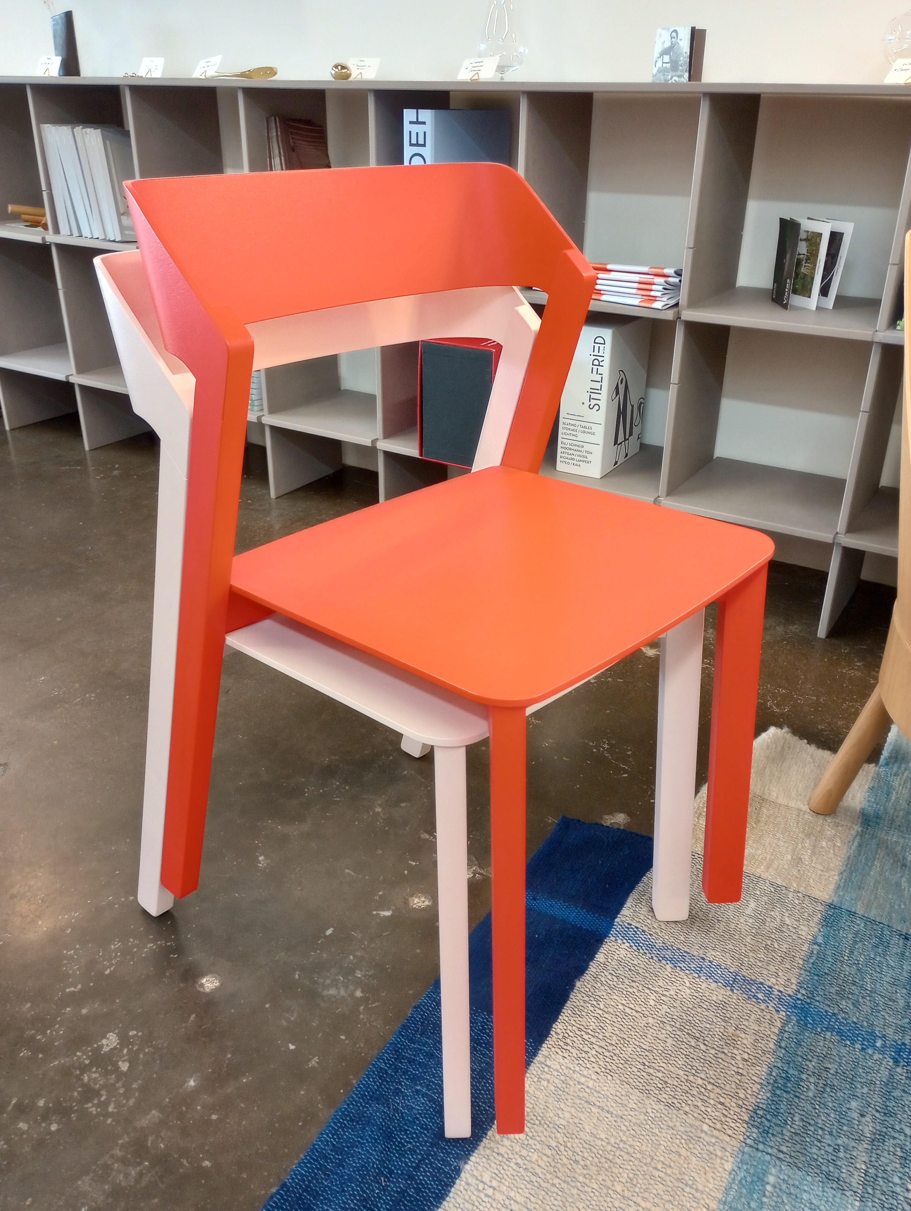 A modern, stackable Chair Merano by TON features a vibrant red seat and backrest made of bent plywood, with white and light brown legs. It complements a room with bookshelves and a blue-and-beige rug.