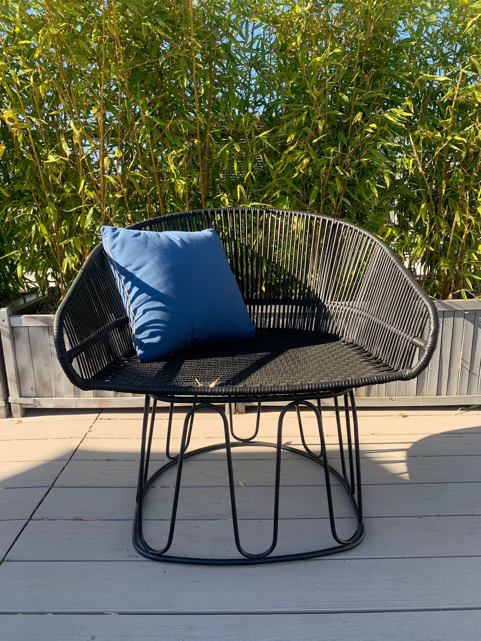 Ames Circo Chairs from the sale collection feature a blue cushion on a black wire-frame design, set elegantly on a wooden deck. Tall green bamboo in the background acts as a natural screen, casting shadows over this exquisite piece of handcrafted furniture and the deck.