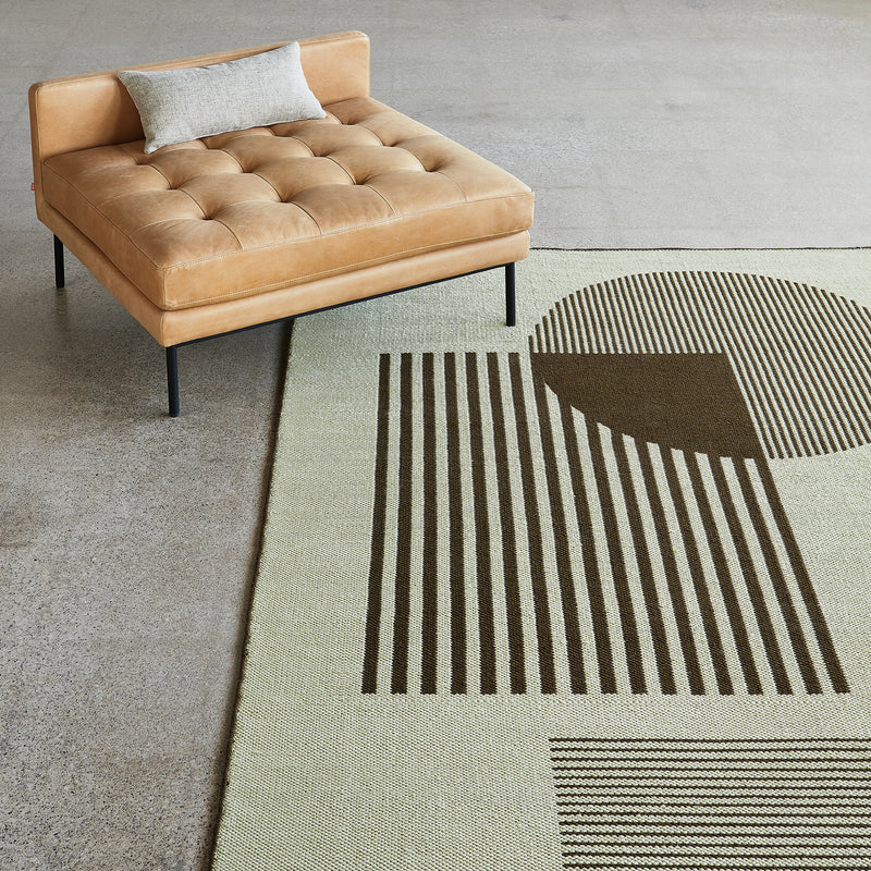 Beige rug with dark brown linear geometric patterns including a large striped circle and rectangle on concrete floor