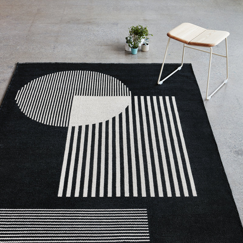 Black rug with large cream geometric shapes including a vertically striped half circle and square on concrete floor
