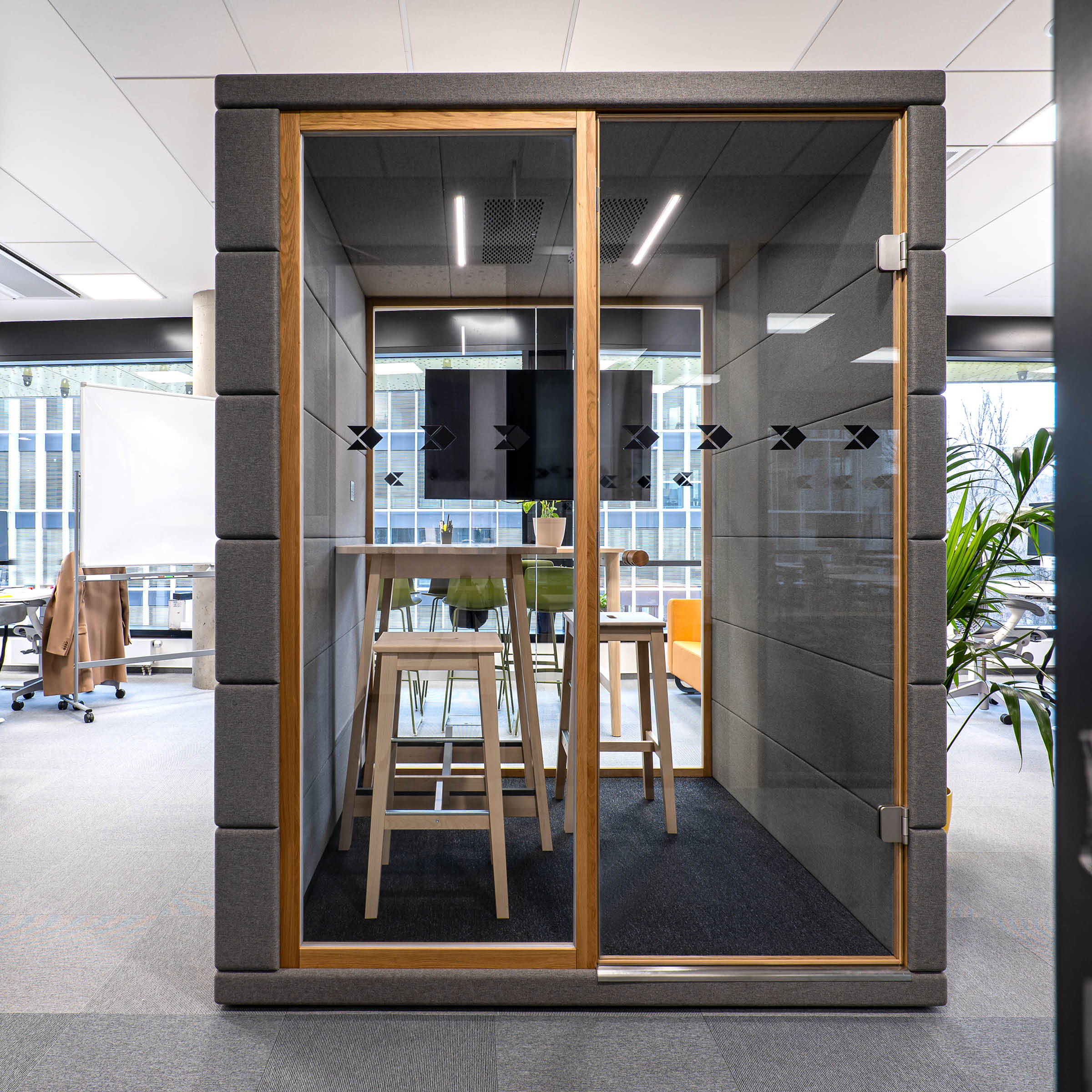 Enhance office privacy with SilentLabs OFFICE POD CUBIQ, a modern workspace featuring glass walls, two high stools, and a screen. Ideal for open offices, its designed to blend seamlessly with nearby desks and plants.