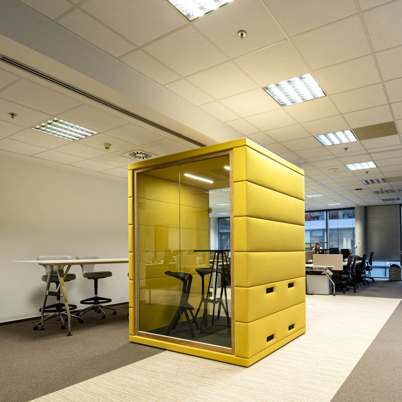 A modern office space features SilentLabs yellow OFFICE POD CUBIQ in the center, surrounded by desks and chairs. The soundproof booth has glass doors for enhanced privacy, with overhead fluorescent lighting illuminating the workspace.