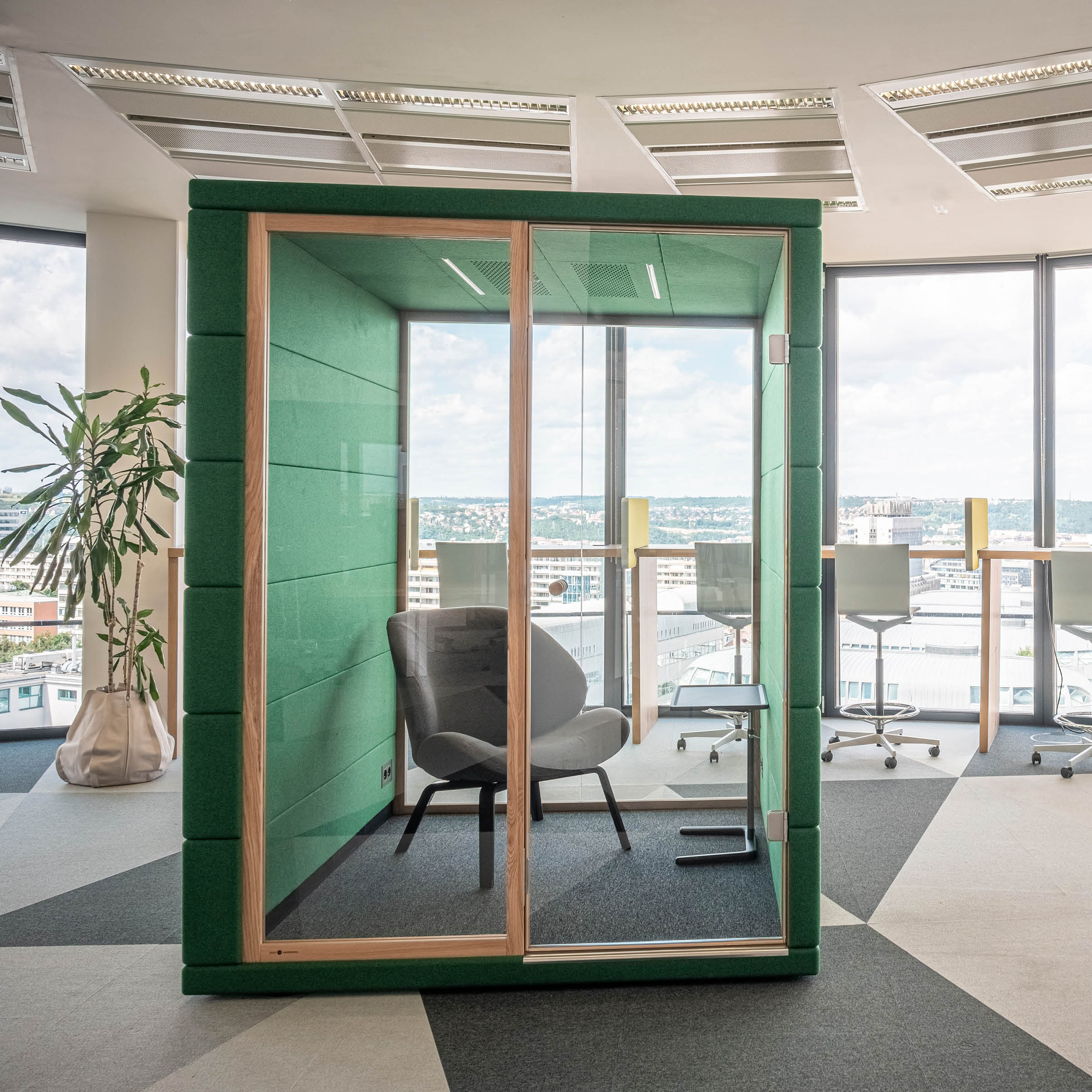 The contemporary workspace features SilentLabs OFFICE POD CUBIQ, a green soundproof phone booth with a glass door and single chair for complete office privacy. Tall windows offer city views, while high tables with stools nearby add functionality.