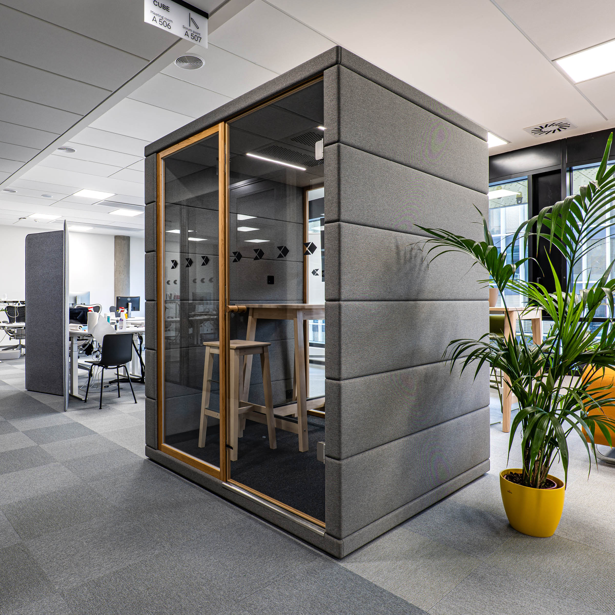 A modern workspace includes a SilentLab OFFICE POD CUBIQ featuring soundproof glass doors, gray padding, and two wooden stools. Its placed beside open workstations and a potted plant in a yellow pot, all under bright overhead lighting for optimal office privacy.