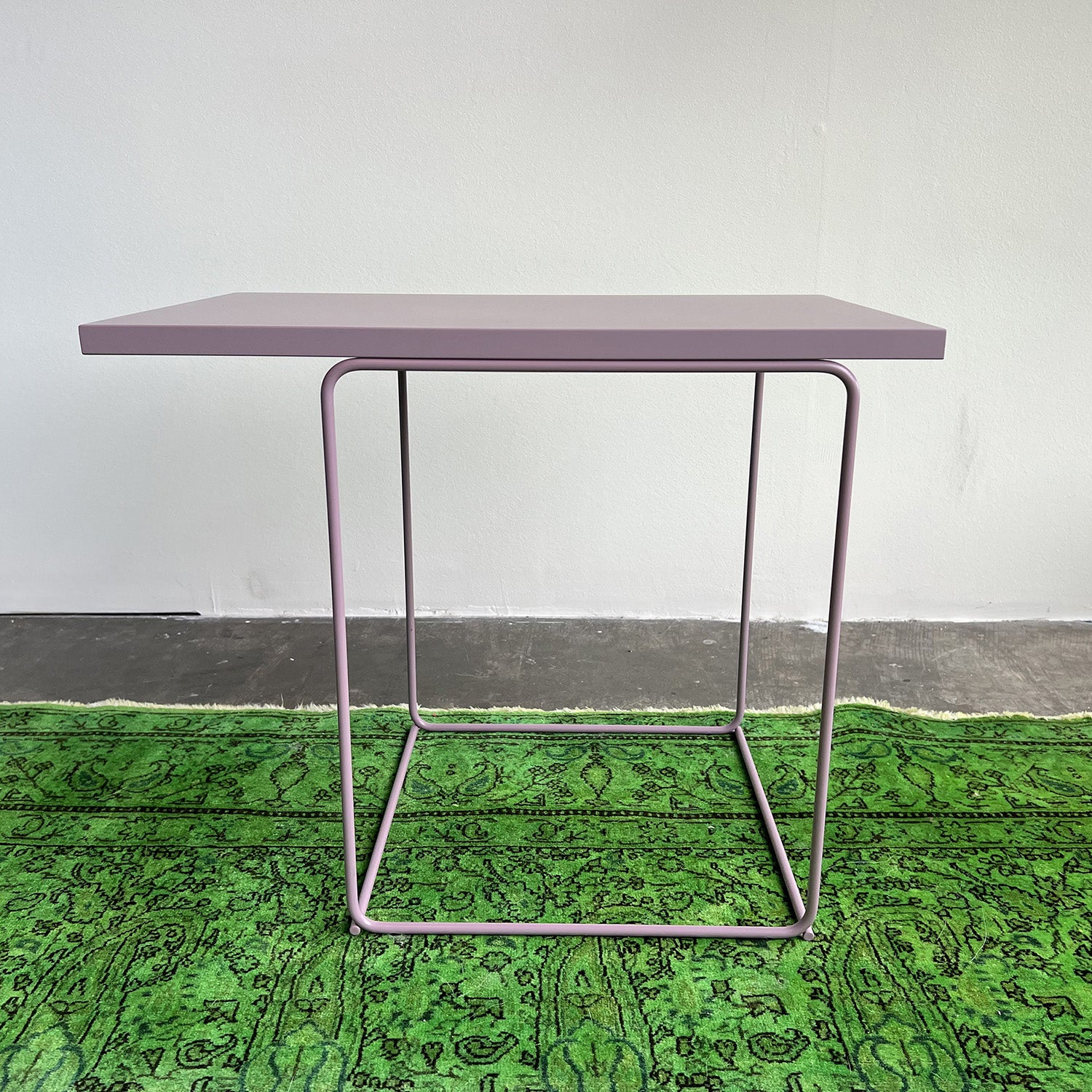 The DL1 Tangram Side Table Asymmetric by Loehr features a minimalist design with thin, light purple metal legs and top. It stands on a vibrant green patterned rug against a plain, light-colored wall.