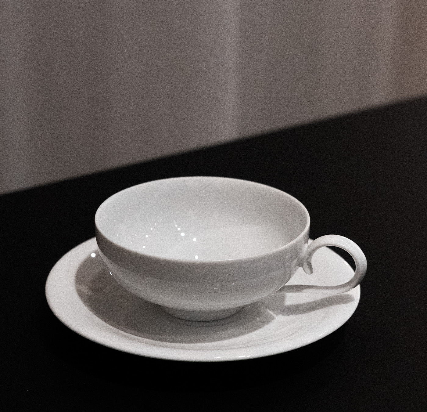 The Augarten - Ena Collection teacup and saucer, featuring a handle and elegant design reminiscent of Vienna Porcelain Manufactory, rests on a black surface with a softly lit, neutral background.