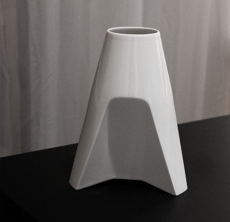The Augarten Rocket Vase by Thomas Feichtner, a uniquely shaped white porcelain piece with a wide base and narrow opening, crafted by the Vienna Porcelain Manufactory, rests elegantly on a dark surface against a light curtain backdrop.