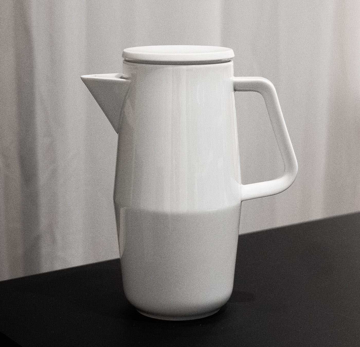 The Augarten Orbit Collection, a white ceramic pitcher with a semi-matte polished rim and lid, showcases rotational shapes. It is displayed on a black surface with light-colored, softly draped fabric in the background.