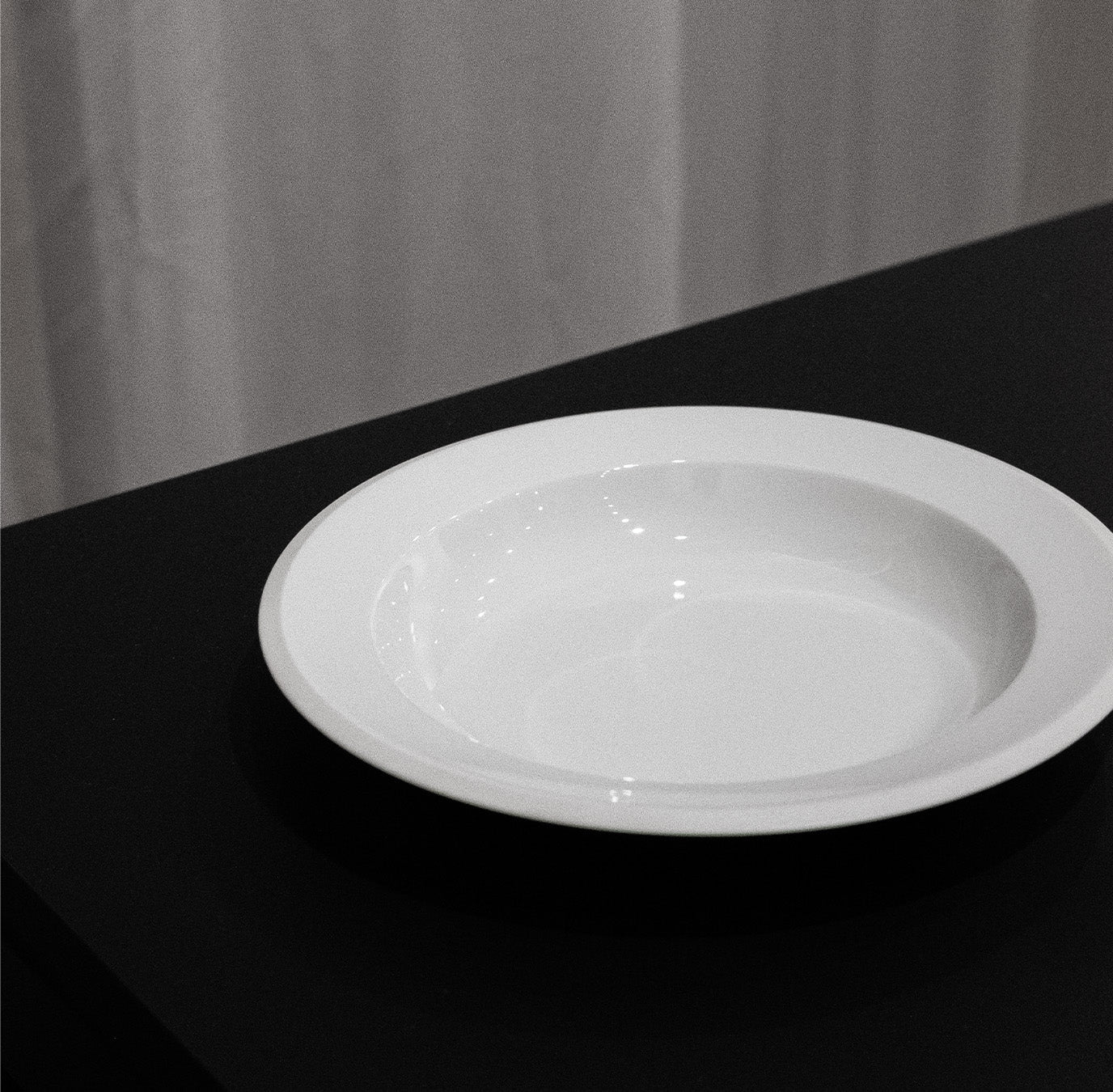 The Augarten Orbit Collection plate, showcasing the minimalist elegance of white porcelain with a semi-matt polished rim, rests on a dark surface. Softly gathered fabric in the background complements this refined Augarten tableware.