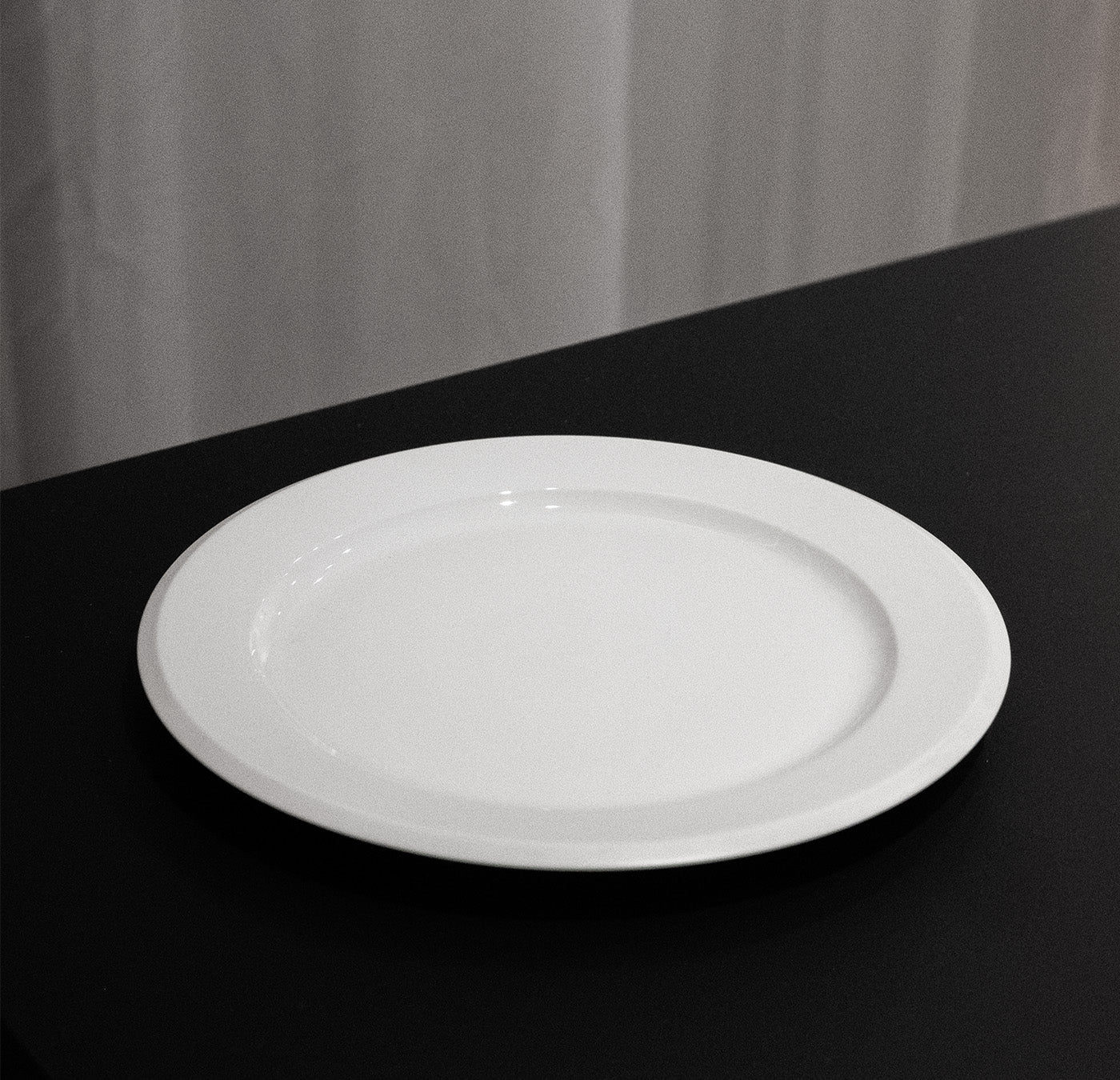 An empty white plate from the Augarten Orbit Collection - Sale, featuring a semi-matt polished rim, sits on a black tabletop against a grey curtain backdrop.