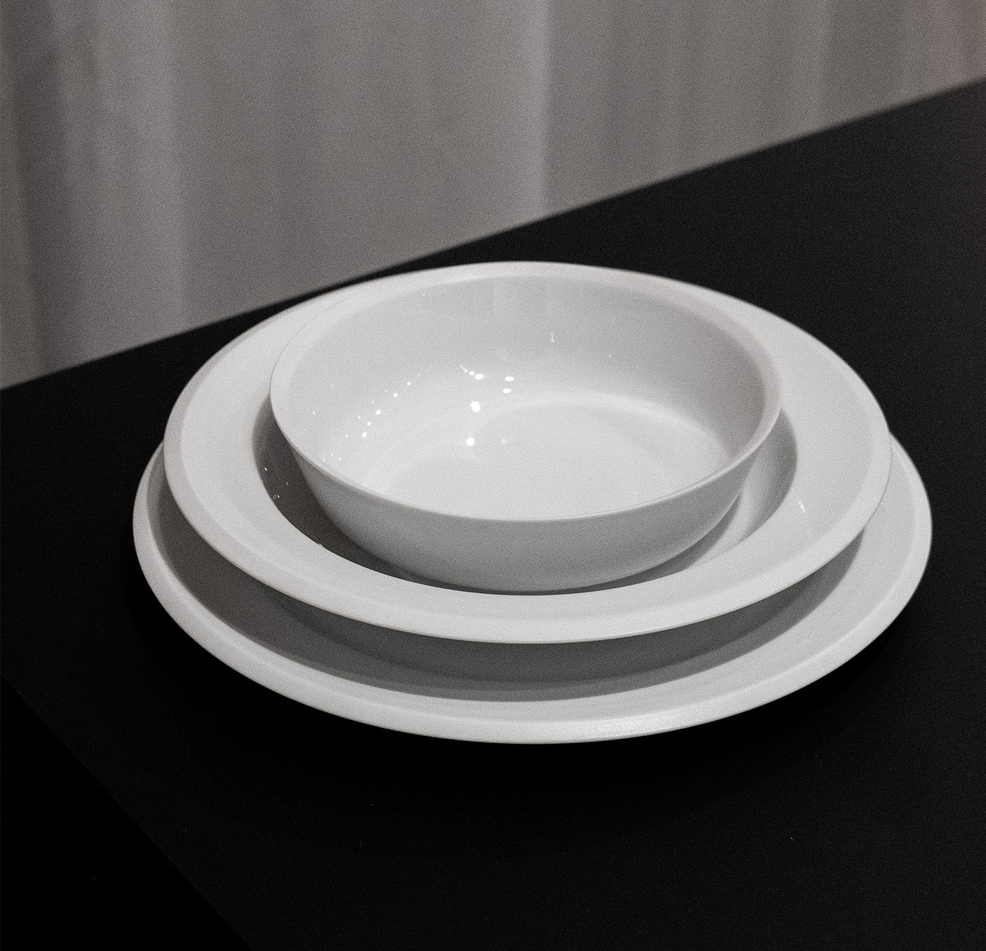 The Augarten Orbit Collection, featuring three porcelain tableware pieces with rotational shapes—a large dinner plate, a smaller plate, and a bowl—rests elegantly on a black surface. The semi-matt polished rim subtly glistens against the backdrop of a white curtain.