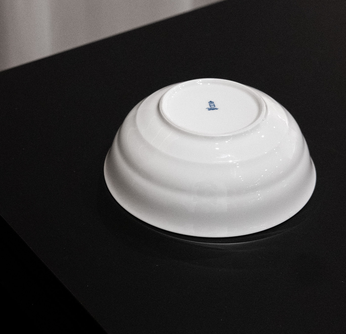 An Augarten Opus Collection ceramic bowl, echoing Austrian design, rests upside down on a black surface, revealing its blue logo underneath.