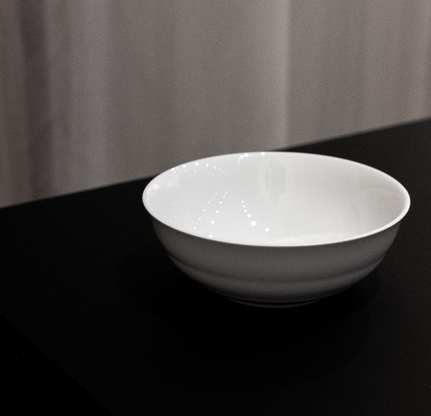 The Augarten Opus Collection bowl, with sleek Art Deco-inspired elegance, rests on a dark surface amid a gently lit gray background, showcasing subtle reflections that enhance its refined beauty.