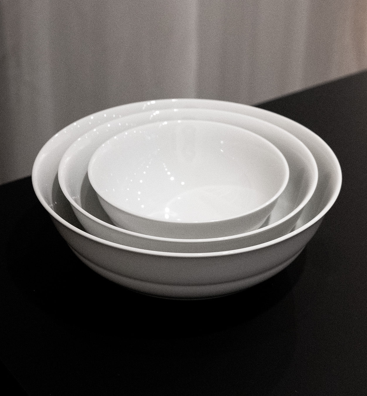 Three white porcelain bowls from the Augarten - Opus Collection are nestled together on a black surface against a light curtain backdrop, perfectly aligned for an aesthetically pleasing arrangement.