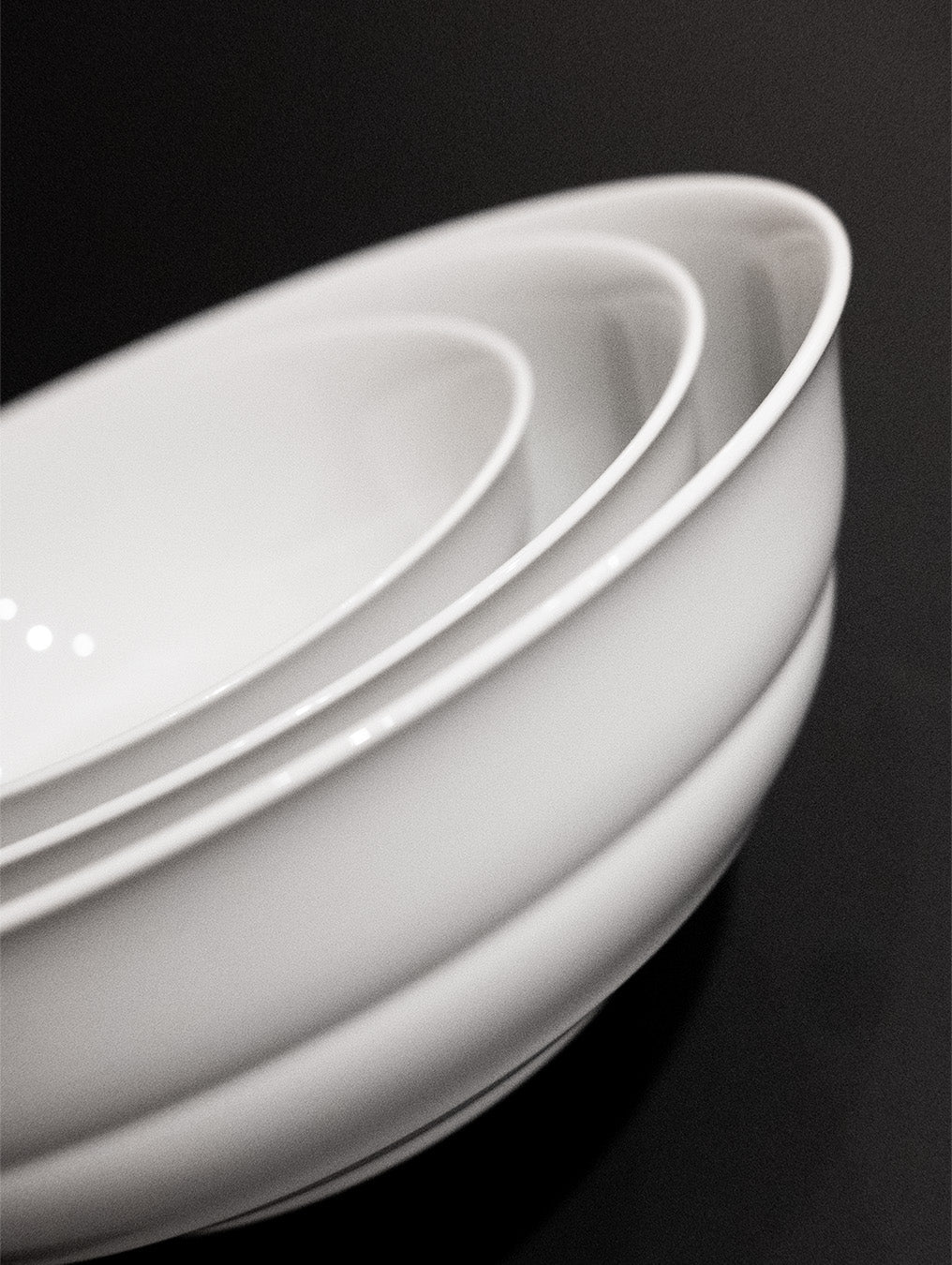 A close-up of four nested white Augarten Opus Collection bowls, stacked elegantly, graces the smooth black surface. Arranged diagonally, they create a striking contrast between the refined Austrian brand ceramics and the dark background.