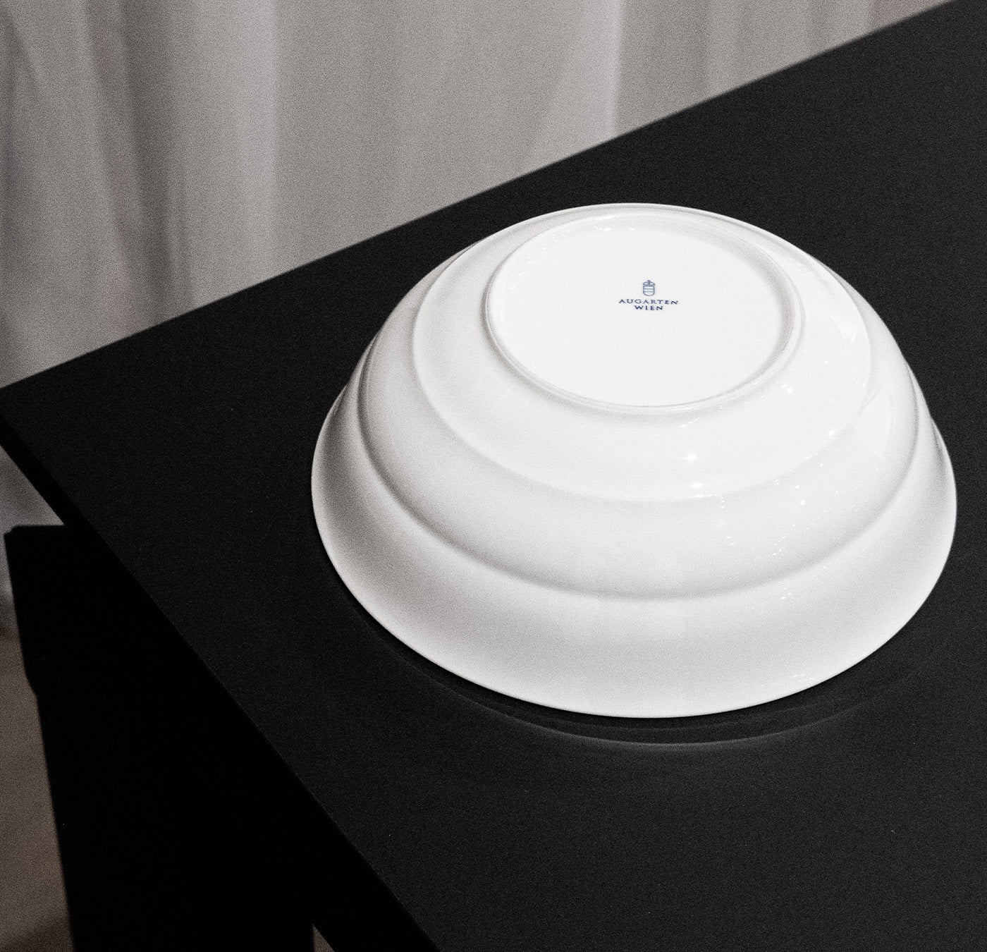 An inverted white ceramic bowl, part of Augartens Opus Collection, sits on a black table. A small blue emblem graces the bottom, enhancing its elegance and nodding to its Austrian ceramics heritage.