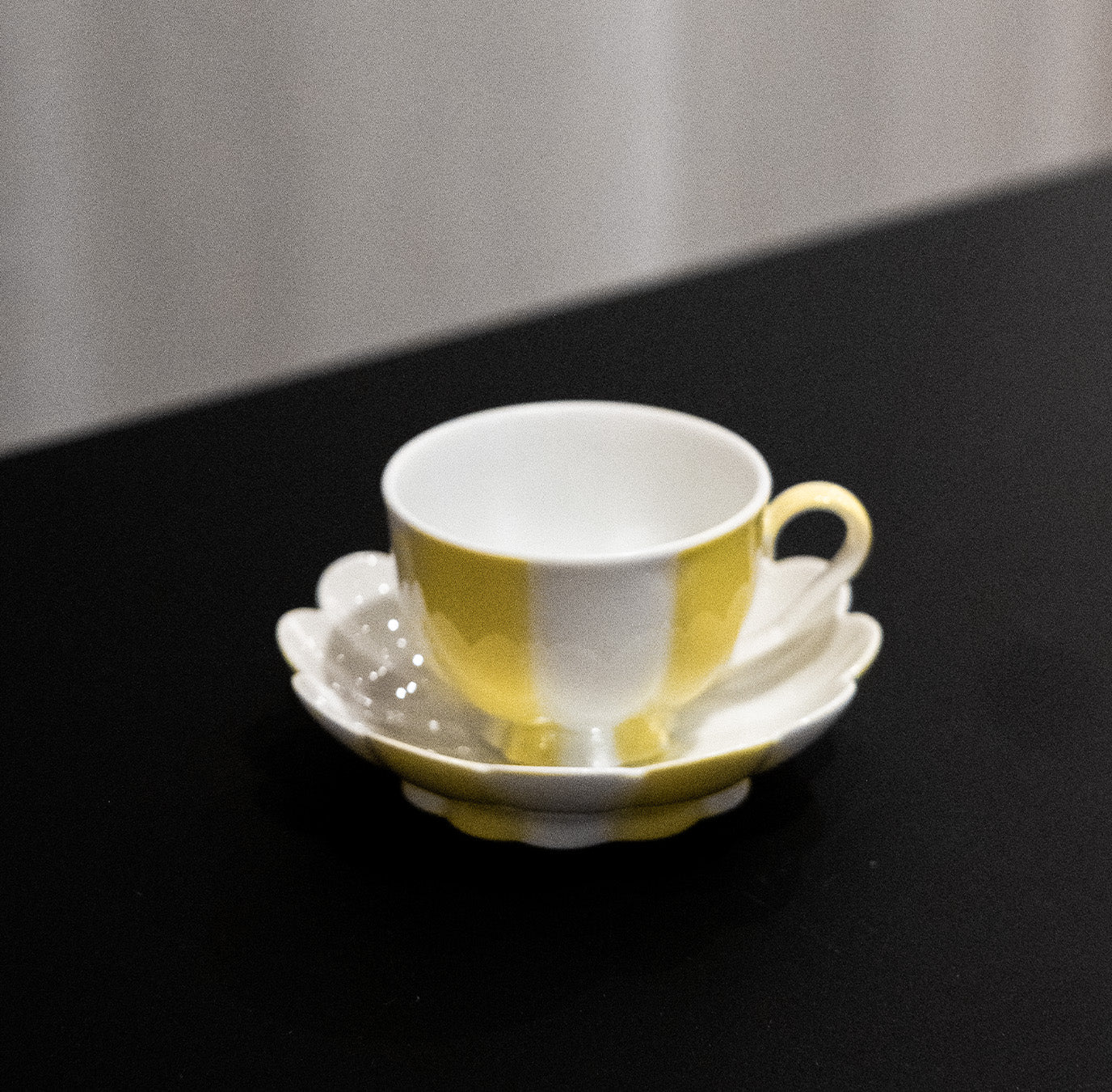 A yellow and white Melon Mocha Cup by Augarten, with a matching saucer and teaspoon, sits on a black surface. The soft lighting casts shadows that highlight the glossy Vienna Porcelain finish of this exquisite piece.