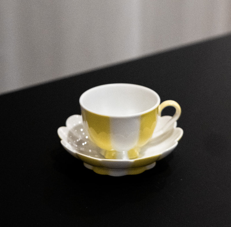 A yellow and white Melon Mocha Cup by Augarten, with a matching saucer and teaspoon, sits on a black surface. The soft lighting casts shadows that highlight the glossy Vienna Porcelain finish of this exquisite piece.