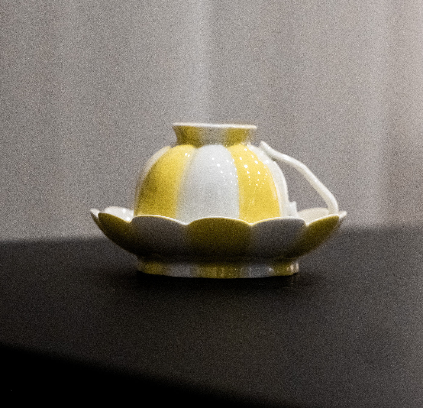 A small, hand-painted ceramic teapot by Augarten, reminiscent of Vienna Porcelain, features a scalloped design and sits on its matching saucer. This yellow and white teapot with a round body and short spout resembles a flower bud resting on petals.