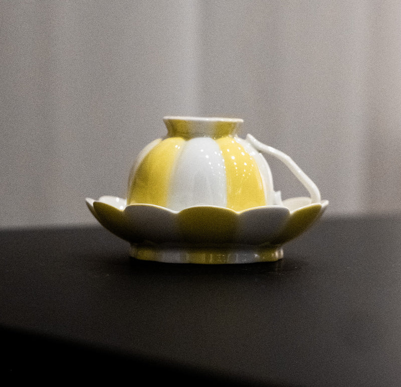 A small, hand-painted ceramic teapot by Augarten, reminiscent of Vienna Porcelain, features a scalloped design and sits on its matching saucer. This yellow and white teapot with a round body and short spout resembles a flower bud resting on petals.
