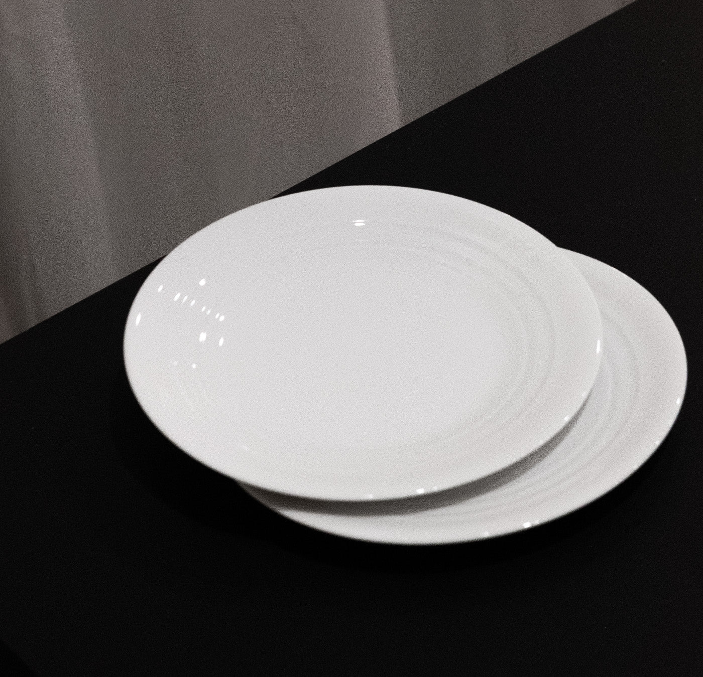 Two stacked plates from the Augarten - Opus Collection rest on a black surface with a curtain backdrop, showcasing a subtle circular ridge design. These elegant Austrian ceramics by Augarten evoke refined craftsmanship.