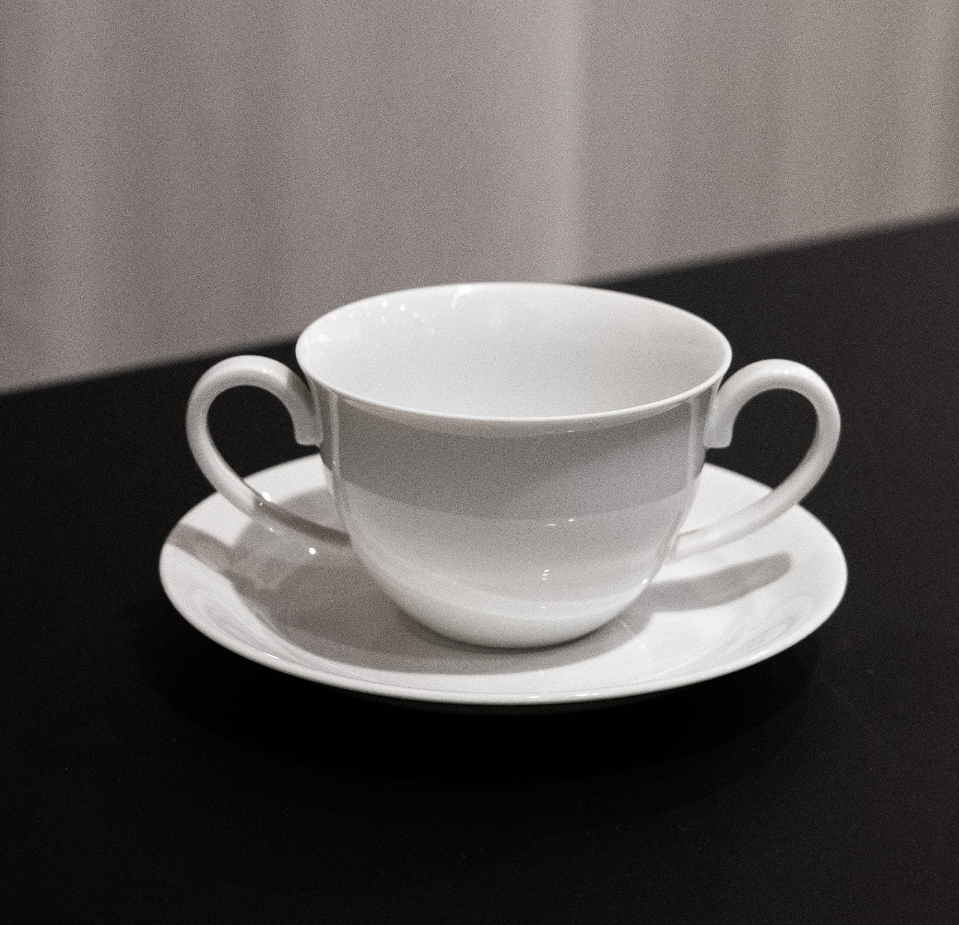 A white, double-handled teacup from the Augarten Opus Collection is on a matching saucer against a black surface. A lightly blurred, light-colored curtain in the background adds elegance to this Art Deco tableware setting.