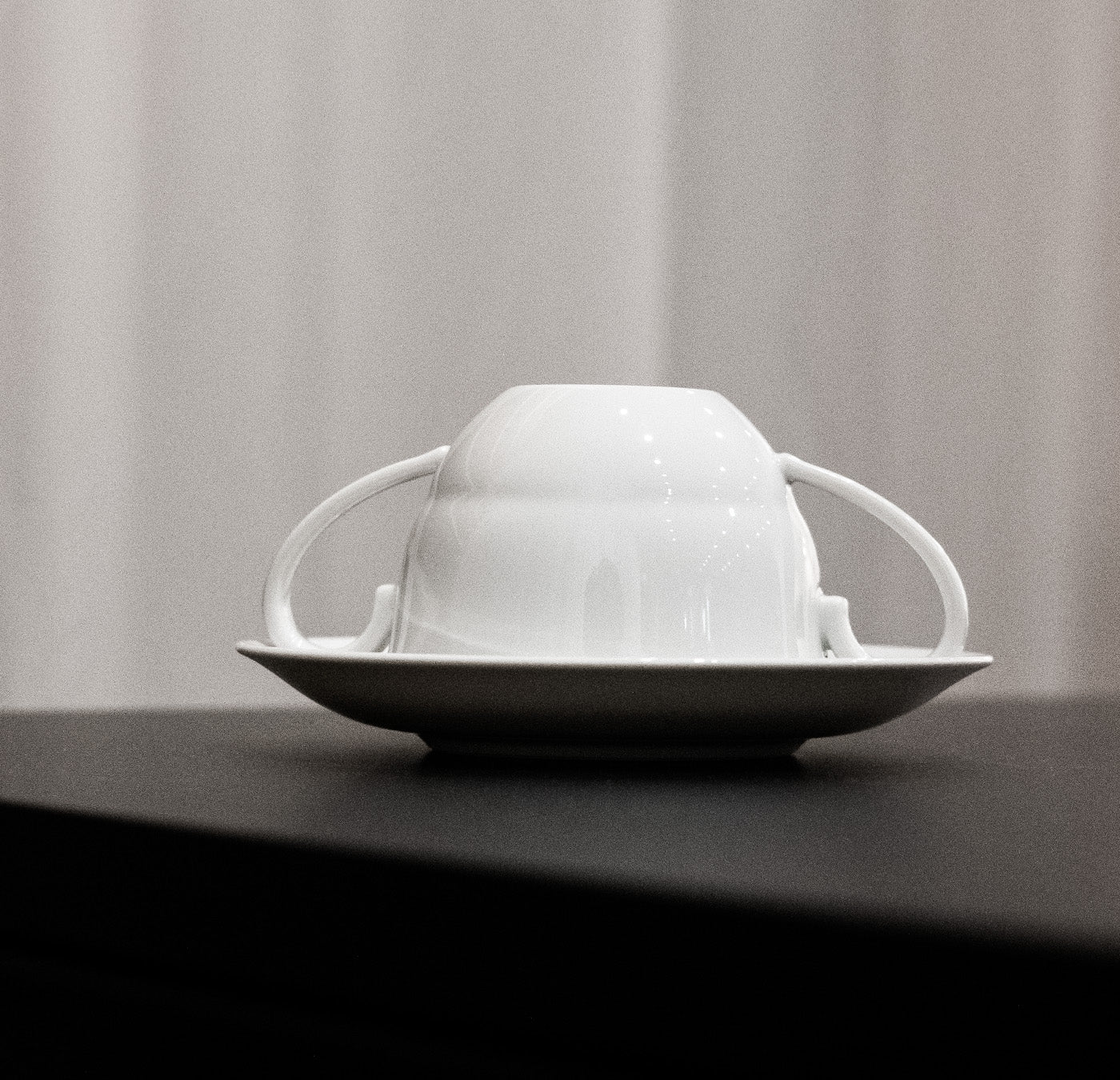 An upside-down Augarten Opus Collection cup with two handles rests on a saucer, placed on a dark surface against a light, blurred background.