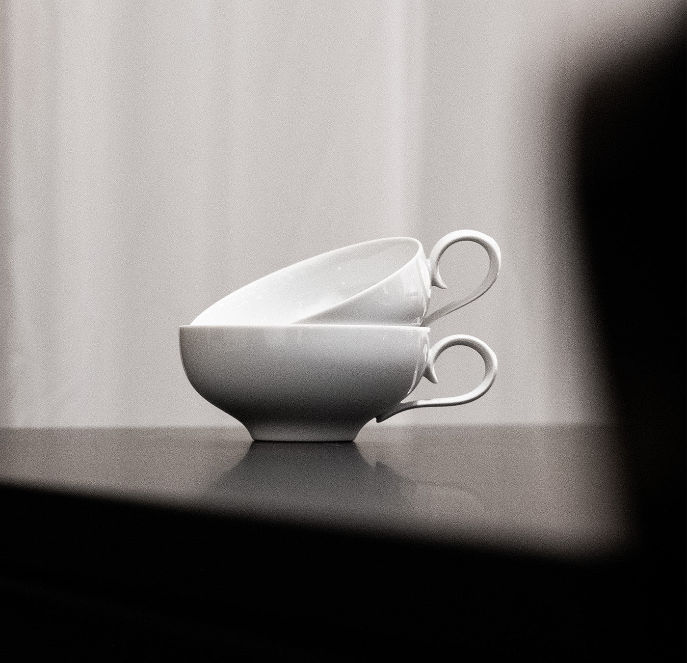 Two white teacups from Augartens Ena Collection are stacked horizontally on a black reflective surface, with a soft, blurred background providing depth.