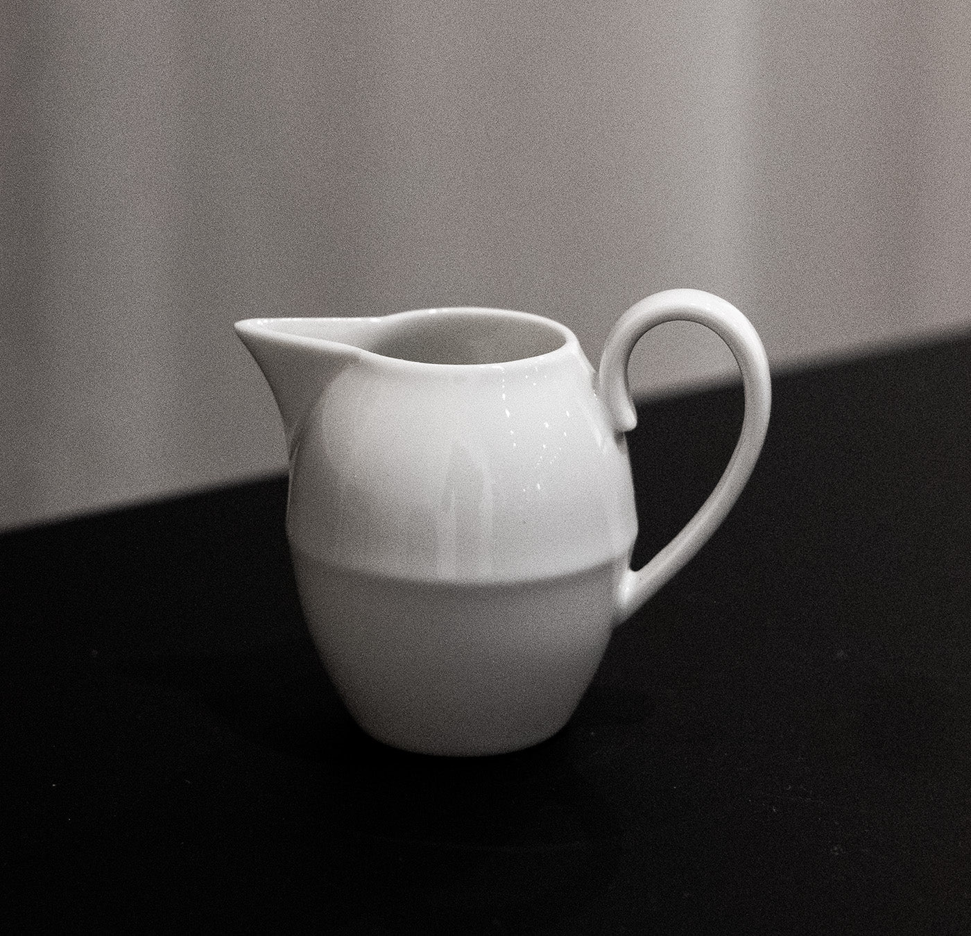 A small, white ceramic pitcher from Augartens Opus Collection sits on a dark surface. With a curved handle, short spout, and round body, it resembles Vienna Porcelain. The design is simple and classic against a plain, slightly blurred background.