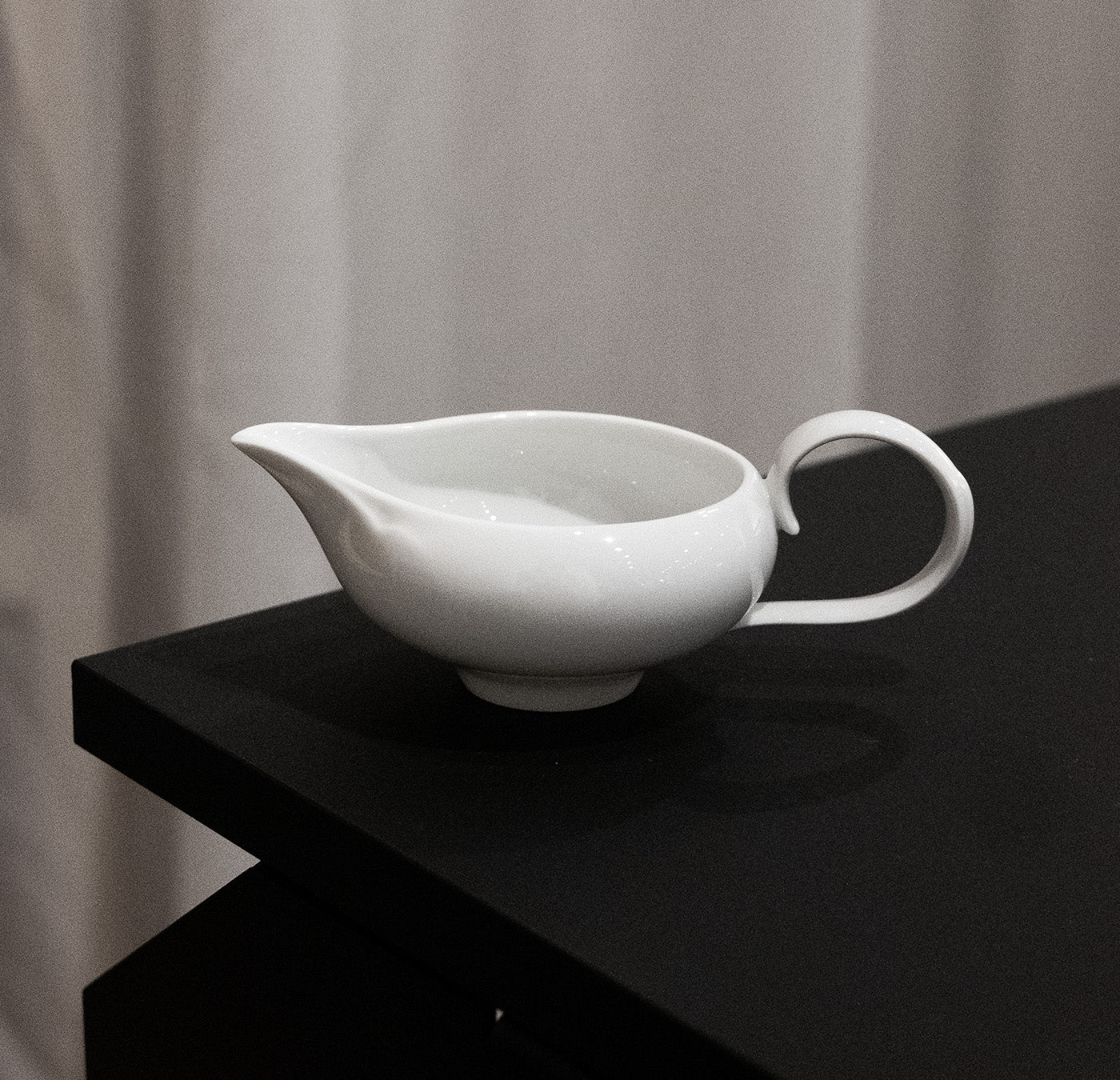 An Augarten Ena Collection small white ceramic pitcher with a curved handle is placed on the corner of a dark table against a light, softly textured background, echoing the artistry of Vienna Porcelain Manufactory.