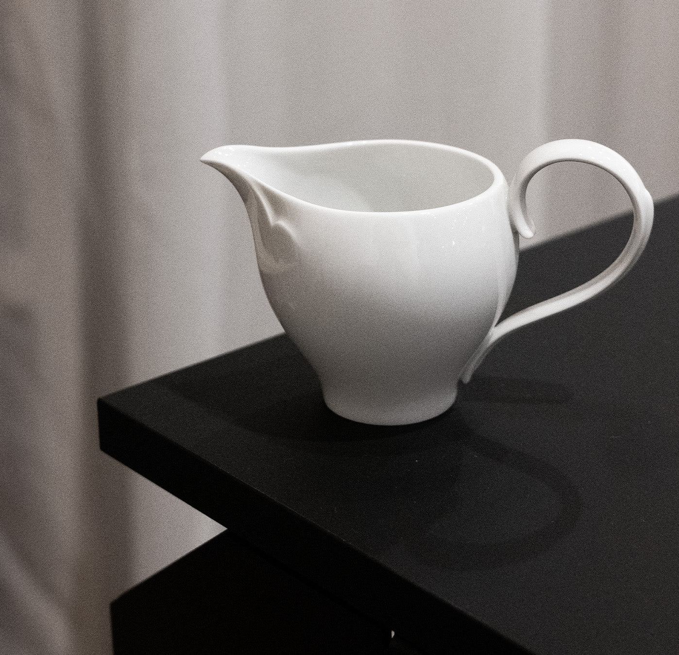 A white porcelain pitcher from the Augarten Ena Collection, featuring a curved handle and spout, is set against a black surface with softly blurred light-colored curtains in the background.
