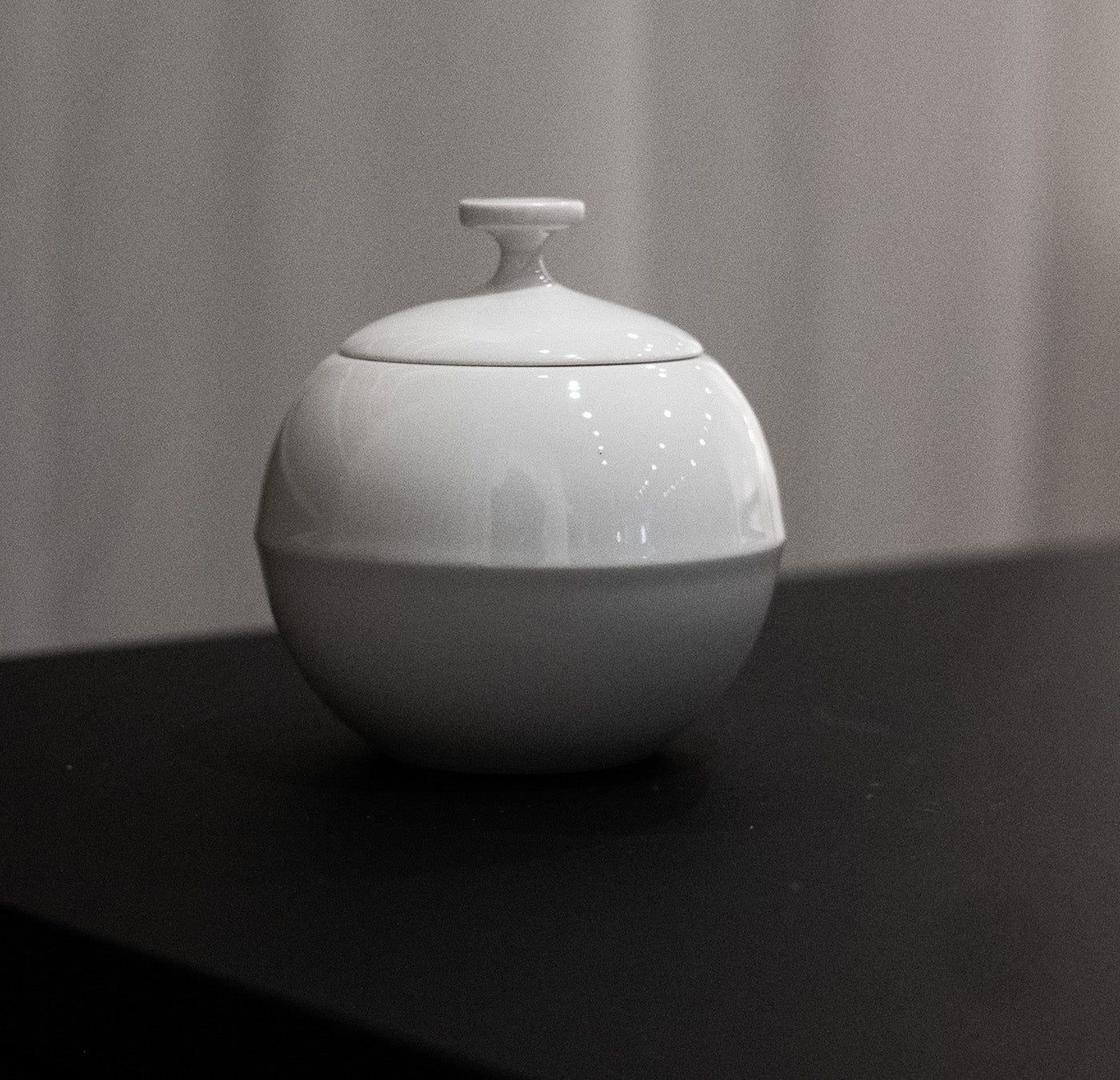 A sleek, round, white ceramic container from the Augarten Opus Collection sale sits on a black surface. Its globe-like design echoes Vienna Porcelain elegance, with softly blurred vertical lines enhancing its modern aesthetic and subtle sophistication.