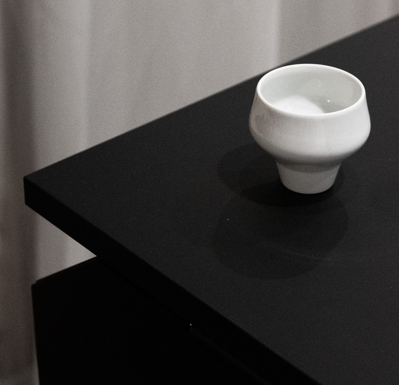 A small porcelain cup from Augartens Shortcut Sale sits on a matte black table, creating a minimalistic scene against a blurred white curtain backdrop. Its soft shadow enhances the multifunctional pieces elegance.