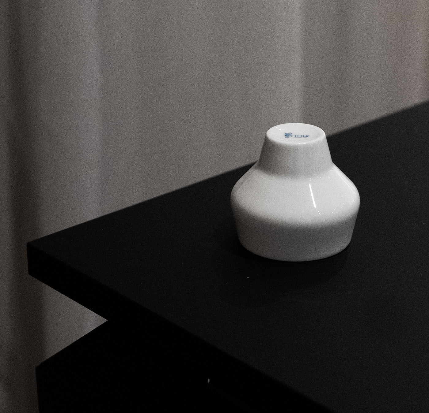 A small white porcelain vase from Augarten sits on a black tabletop, echoing the elegance of Vienna Porcelain Manufactory. A neutral draped curtain acts as the background.