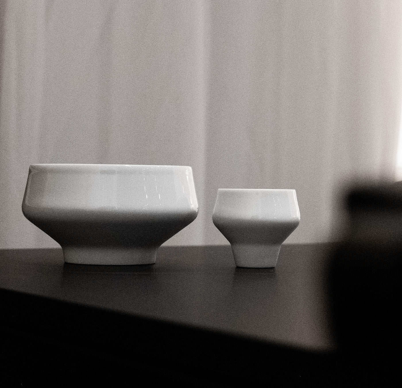 Two white Augarten ceramic bowls, of different sizes, sit elegantly on a dark surface. The larger bowl is on the left, and the smaller on the right. Soft, light-colored curtains in the background create a minimalist setting.