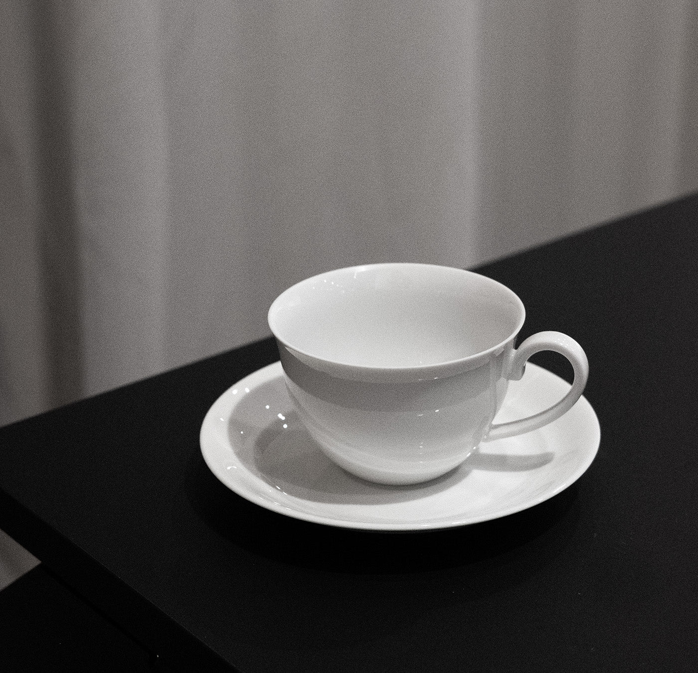 An Augarten - Opus Collection teacup, evoking classic Vienna Porcelain, rests gracefully on its matching saucer on a dark table, with a light curtain elegantly draped in the background.