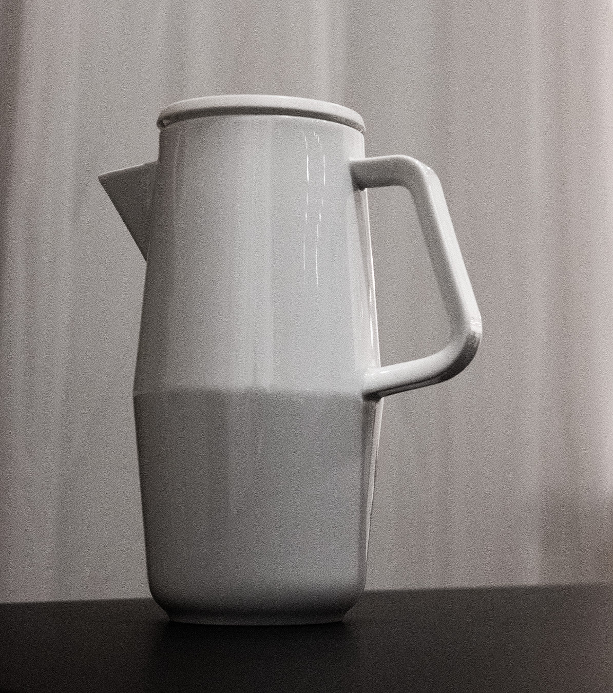 A black and white image features the Augarten Orbit Collection pitcher with a lid and angular handle, showcasing its semi-matt polished rim on a dark surface. A light curtain in the background emphasizes the elegance of this Augarten porcelain tableware piece.
