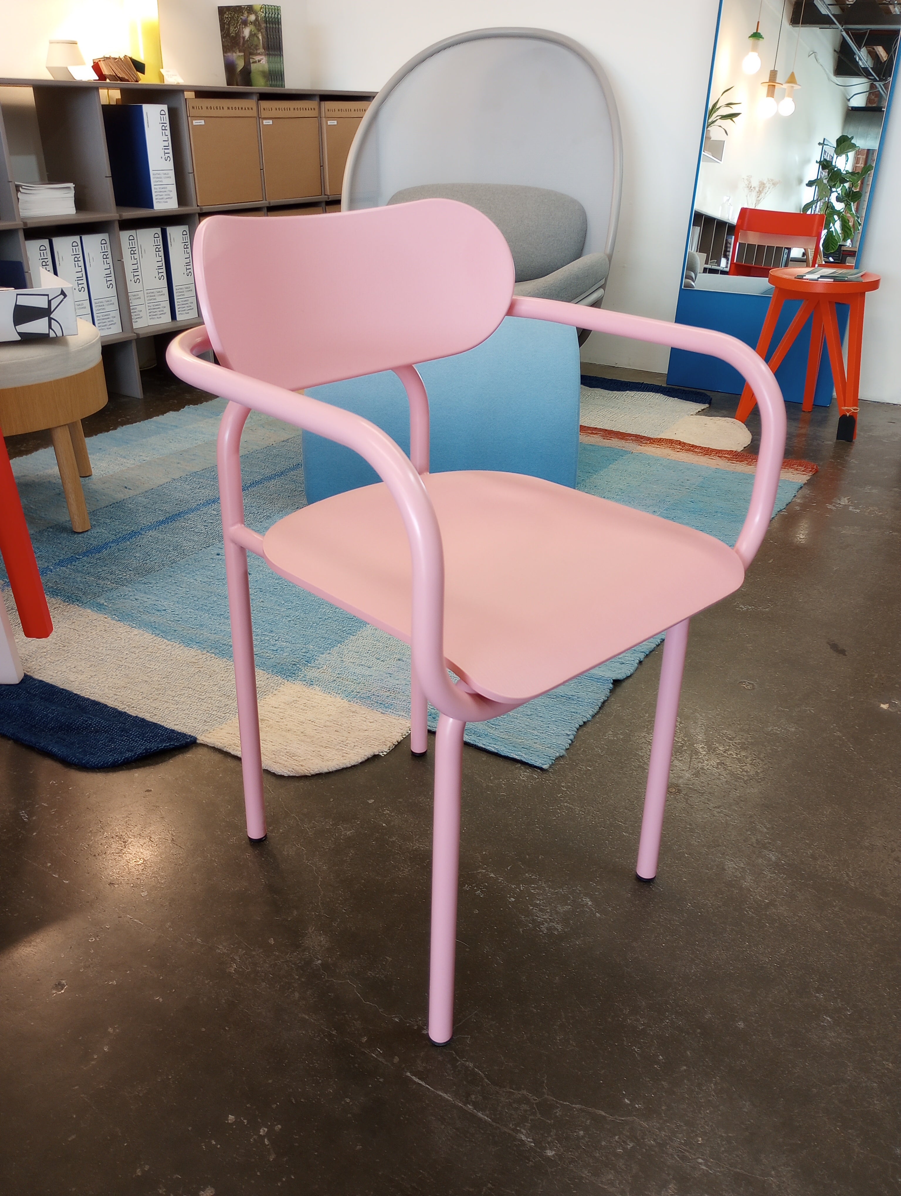 A pink Double U Armchair - Sale by Victor Foxtrot with a rounded backrest and armrests sits on a gray floor. The backdrop showcases striking aesthetics, including a colorful rug, shelves with binders, a sofa, and modern decor like orange tables and plants.